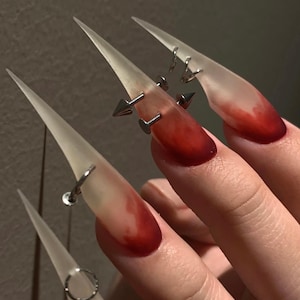 May include: Long, pointed, translucent off-white artificial nails featuring a gradient red color at the tips. The nails are decorated with various silver-toned metal piercings, including small hoops, flat-back studs, and sharp conical spikes.