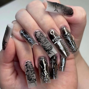 Pode incluir: Unhas acrílicas longas e quadradas com um gradiente escuro e esfumaçado. Algumas unhas apresentam intrincados desenhos de linhas pretas, enquanto outras têm pingentes de adaga prateada e ornamentos prateados ornamentados. O design geral é gótico e ousado.