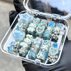 Op de afbeelding: Een doorzichtige plastic doos gevuld met een set nepnagels. De nagels hebben verschillende ontwerpen in blauwe, zilveren glitter- en wittinten. Sommige nagels hebben ster-, hart- en strikaccenten en sommige hebben een cartoonkat.