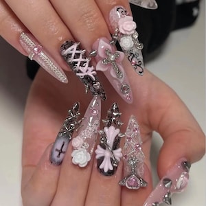 May include: Long stiletto-shaped artificial nails featuring a complex design with pink and white 3D roses, silver cross charms, black lace patterns, metallic studs, and pink rhinestones. The nails incorporate corset-style lacing details.