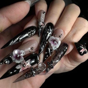May include: A set of long, pointed acrylic nails. The nails are painted black, nude, and clear, with silver accents, floral designs, and text. Some nails feature 3D flower embellishments and metal charms.