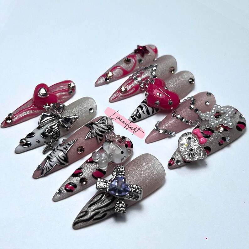 Op de afbeelding: Een verzameling lange, puntige kunstnagels met verschillende ontwerpen. Sommige nagels hebben zebra strepen, luipaardprint en glitter. Andere hebben hart-, strik- en edelsteenversieringen. De nagels zijn in tinten roze, zilver en wit.