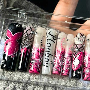 May include: A set of long, acrylic nails with various designs. The nails feature a combination of black, white, and pink colors, with accents like rhinestones, a crown, and the word "Playboy". Some nails have a zebra stripe pattern.