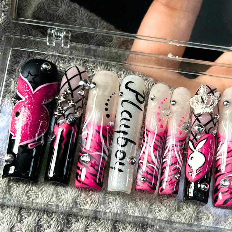 May include: A set of long, acrylic nails with various designs. The nails feature a combination of black, white, and pink colors, with accents like rhinestones, a crown, and the word "Playboy". Some nails have a zebra stripe pattern.