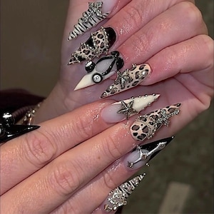May include: A set of long, pointed acrylic nails with various designs. The nails feature leopard print, zebra stripes, and embellishments like stars, rhinestones, and a pool ball. Some nails are black and white, while others are nude and beige.