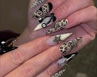 Op de afbeelding: Een set lange, puntige acrylnagels met diverse ontwerpen. De nagels hebben luipaardprint, zebra strepen en versieringen zoals sterren, strass en een poolbal. Sommige nagels zijn zwart-wit, andere nude en beige.