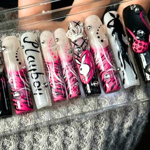 May include: A set of long, artificial nails with a variety of designs. The nails feature a combination of pink, black, and white colors, with zebra stripes, the word "Playboy", and a Playboy bunny logo. Some nails are adorned with rhinestones and a crown.