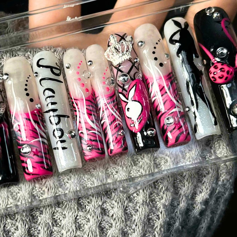 May include: A set of long, artificial nails with a variety of designs. The nails feature a combination of pink, black, and white colors, with zebra stripes, the word "Playboy", and a Playboy bunny logo. Some nails are adorned with rhinestones and a crown.
