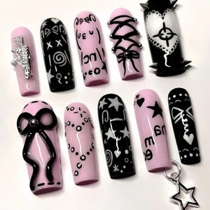 Puede incluir: Un conjunto de diez uñas artificiales con varios diseños. Las uñas son principalmente rosas y negras, con adornos que incluyen lazos, estrellas y texto. Algunas uñas presentan dijes y tachuelas plateadas. La estética general es atrevida y elegante.