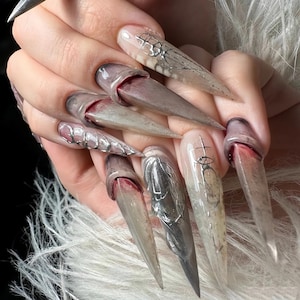 May include: A set of long, pointed acrylic nails with a unique design. The nails feature a gradient of colors, from clear to gray and brown, with silver accents and a blood-red effect at the base. Some nails have intricate silver wire designs.