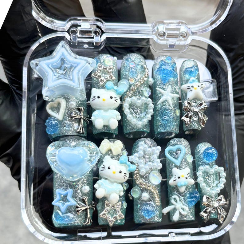 Op de afbeelding: Een set opzetnagels in een doorzichtige plastic doos. De nagels zijn lichtblauw en zilverglitter, met diverse versieringen, waaronder ster-, hart- en strikvormige bedels en Hello Kitty-figuren.
