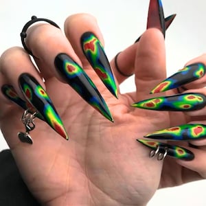 Puede incluir: Uñas largas negras de estilo stiletto con un diseño de mapa de calor en rojo, amarillo, verde y azul. Las uñas están adornadas con anillos plateados y un anillo negro en un dedo. El efecto general es audaz y llamativo.