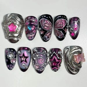 May include: A set of ten artificial nails with various designs. The nails feature silver and pink star and butterfly accents, along with glitter and gem embellishments. The color palette includes black, pink, purple, and silver, creating a glamorous aesthetic.
