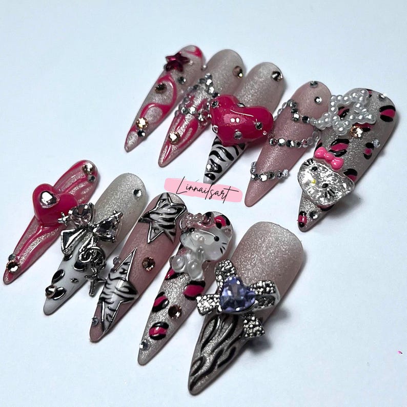 Op de afbeelding: Een verzameling lange, puntige kunstnagels met diverse ontwerpen. Sommige nagels hebben roze en zilveren glitter, hartvormige versieringen en zebra strepen. Andere hebben zilveren strikken, strass en een Hello Kitty-bedel. De nagels zijn gerangschikt op een wit oppervlak.