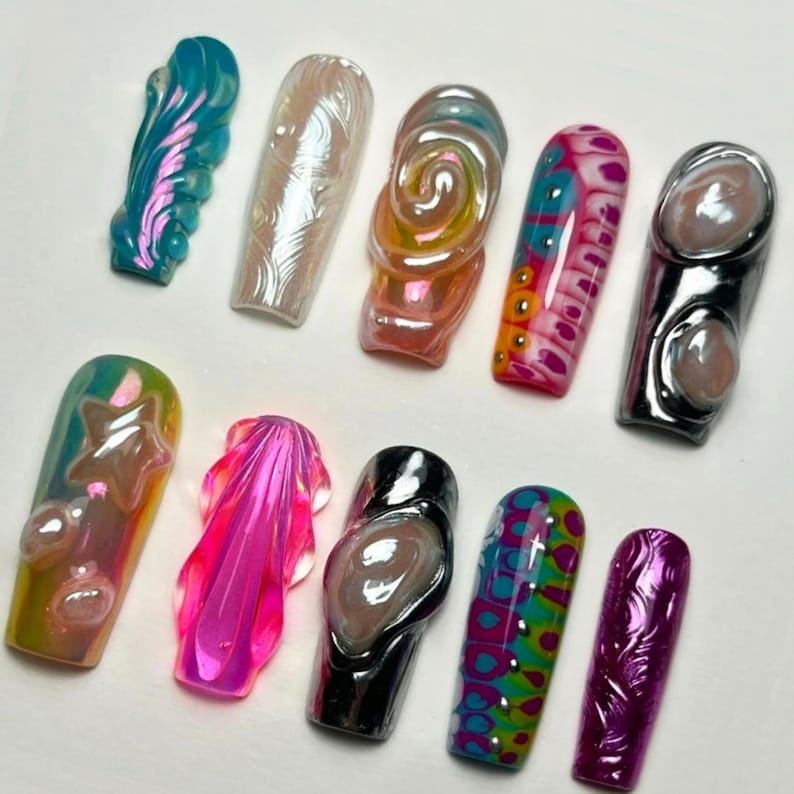 May include: A collection of ten artificial nails in various designs and colors. The nails feature iridescent finishes, swirls, and abstract patterns. Colors include pink, blue, green, and gold. Some nails have 3D elements.