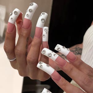 Op de afbeelding: Lange, vierkante kunstnagels in een French manicure-stijl met een nude basis en ondoorzichtige witte uiteinden. Elk wit uiteinde is versierd met meerdere heldere, ronde, gefacetteerde strass-steentjes van verschillende groottes over het oppervlak.