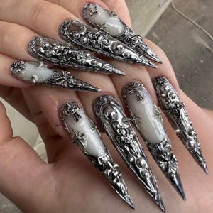 Peut inclure: Ongles allongés, argentés, avec des motifs complexes et ornés. Chaque ongle présente un accent blanc de forme ovale et des détails en métal, notamment des croix, des étoiles et des motifs floraux. Les ongles sont pointus.