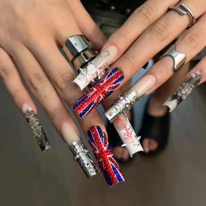 Puede incluir: Uñas postizas largas de forma cuadrada con diversos diseños, incluyendo un patrón de bandera Union Jack en rojo, blanco y azul hecho de pedrería, superposiciones de encaje negro, detalles metálicos plateados y texto rojo en cursiva sobre una base nude.