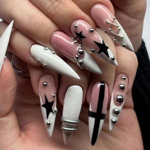 Peut inclure: Ensemble de longs ongles en acrylique pointus avec divers motifs. Certains ongles sont blancs, d'autres roses avec des pointes blanches. Étoiles noires et croix, clous argentés et design argenté lacé.