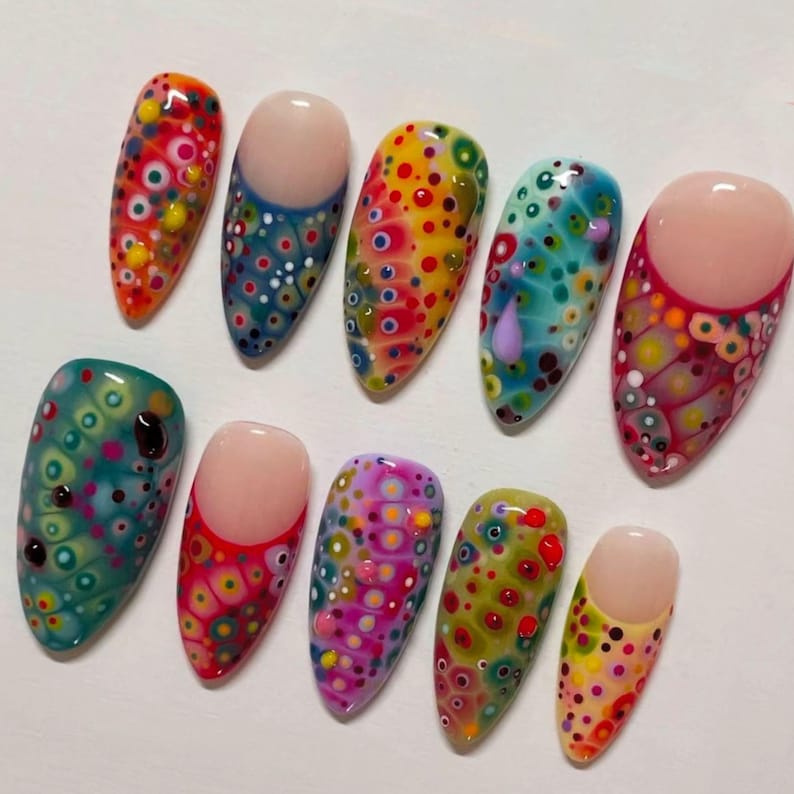 May include: A collection of ten artificial nails with a glossy finish. Each nail features a unique design of colorful, abstract patterns with a variety of dots and circles in shades of red, yellow, blue, and green. Some nails have a nude base.