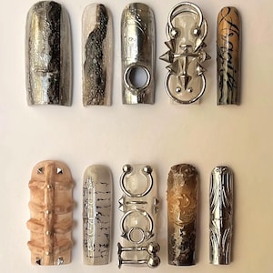 Könnte beinhalten: Eine Sammlung von zehn künstlichen Nägeln mit verschiedenen Designs. Die Nägel weisen metallische, strukturierte und gemusterte Elemente auf, darunter Spikes, Wirbel und verzierte Details. Farben sind Silber, Braun und Transparent.