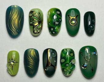 May include: A collection of ten oval-shaped, manicured nails. The nails feature various designs, including gold swirls, a butterfly, and abstract patterns in shades of green, teal, and gold. Some nails have embellishments like gems and bubbles.