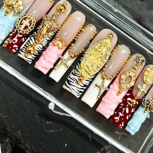 Puede incluir: Un conjunto de uñas artificiales largas y adornadas. Los diseños incluyen cruces doradas, amuletos e iconografía religiosa. Los colores de las uñas incluyen blanco, rosa, estampado de cebra y burdeos. Las uñas se muestran en un recipiente transparente.