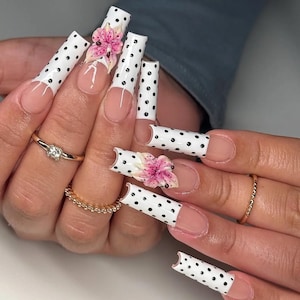 Könnte beinhalten: Lange, quadratische Kunstnägel mit weißer Basis und schwarzen Tupfen. Einige Nägel weisen ein rosa Blumendesign auf. Die Nägel haben ein French-Tip-Design. Goldene Ringe schmücken die Finger.
