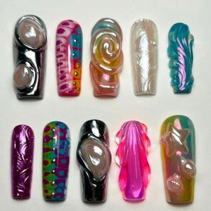 May include: An assortment of ten iridescent, sculpted acrylic nails. Designs include swirls, abstract patterns, and 3D embellishments. Colors range from pink, blue, and green to silver and gold, with a glossy finish. Each nail has a unique shape and design.