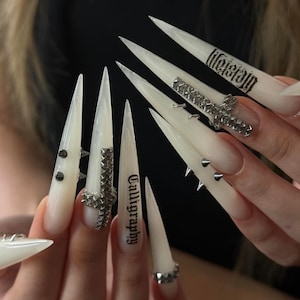 May include: Long, pointed, off-white acrylic nails featuring silver-toned metal embellishments, including small spikes, pyramid-studded cross charms, and black text reading "Calligraphy" and "Lifeless" printed on the surface of the nail extensions.