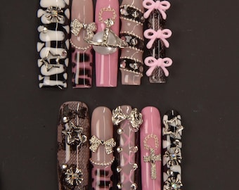 Op de afbeelding: Een set lange, kunstmatige nagels in verschillende ontwerpen. De nagels tonen een mix van zwart, wit, roze en nude. Versieringen zijn onder meer strikken, sterren en decoratieve juwelen. Sommige nagels hebben ruitpatronen en kanten details.