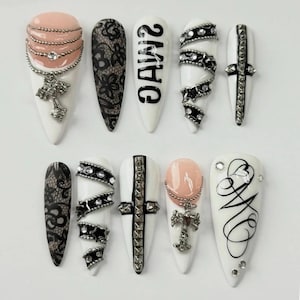 May include: A collection of ten long, white artificial nails with various designs. Some nails feature black lace patterns, silver chains, and cross charms. One nail has the text "2MHC" in black. Other nails have silver and black embellishments.