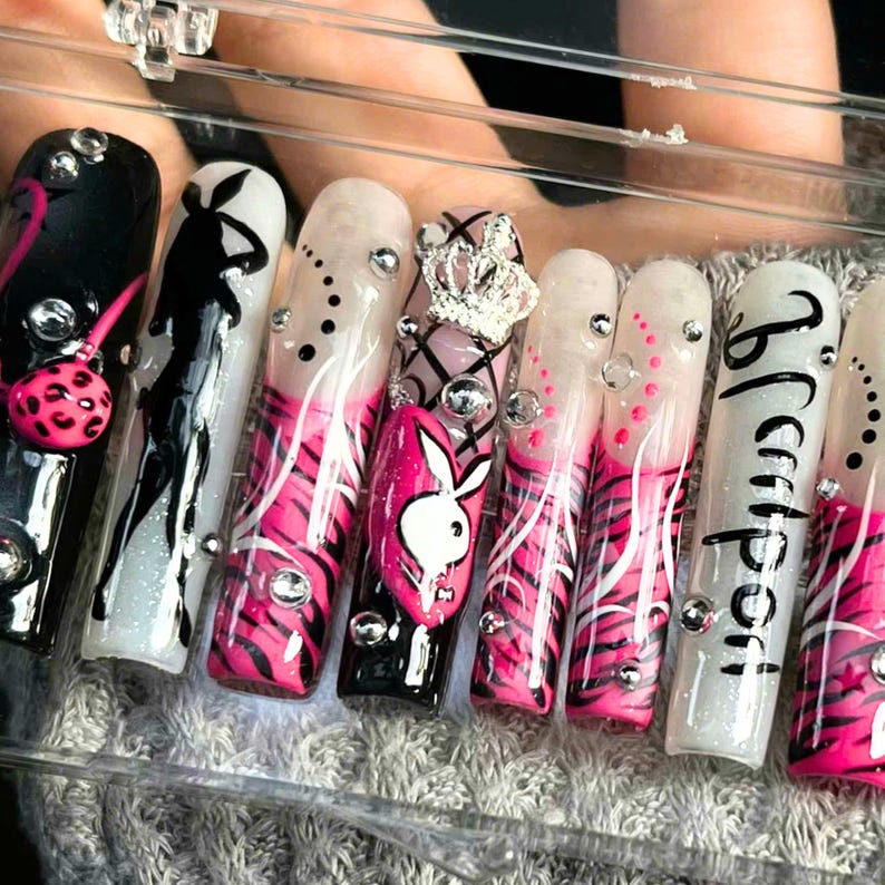 May include: A set of long, artificial nails with various designs. The nails feature black, white, and pink patterns, including a Playboy bunny logo, zebra stripes, and rhinestones. Some nails have text and a crown.