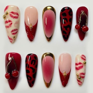 May include: A collection of ten artificial nails with various designs. The nails feature a range of colors, including red, pink, and gold. Some nails have lip prints, leopard spots, or cherry accents. Others have gold trim or star details.