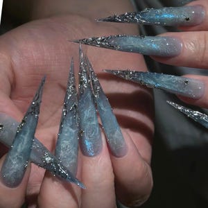 May include: Long, stiletto-shaped artificial nails featuring a gradient of blue and grey tones with a marbled texture. The nails are decorated with intricate, raised silver metallic accents and small, clear embedded rhinestones.