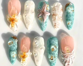 Puede incluir: Una colección de diez uñas artificiales largas en forma de almendra. Las uñas presentan varios diseños, incluyendo una estrella de mar, conchas marinas y acentos florales. Los colores incluyen tonos de rosa, blanco, verde azulado y dorado. Las uñas están dispuestas sobre una superficie blanca.