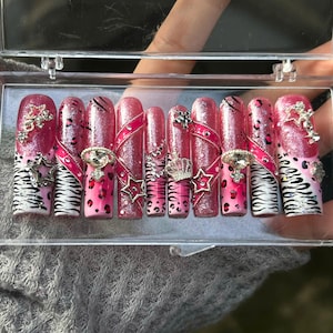 Puede incluir: Un conjunto de uñas postizas rosas y brillantes con varios diseños. Algunas uñas presentan rayas de cebra, estampado de leopardo y pedrería. Otras tienen adornos de estrellas y coronas. Las uñas se muestran en una caja de plástico transparente.