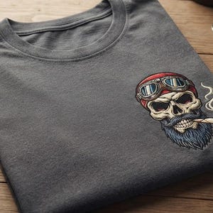 May include: A charcoal gray t-shirt featuring an embroidered graphic of a skull wearing red aviator goggles and a blue beard, with a lit cigarette in its mouth. The text PREMIUM BIKER EMBROIDERY is visible in the bottom right corner of the image.