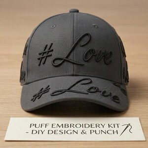 May include: A dark grey baseball cap featuring black raised puff embroidery of the text #Love on the front and brim. A white rectangular card is placed in front of the cap with the text PUFF EMBROIDERY KIT - DIY DESIGN & PUNCH printed on it.