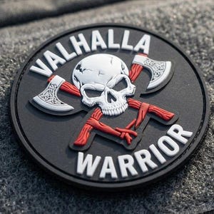Op de afbeelding: Een ronde zwarte tactische patch met een witte schedel gecentreerd over twee gekruiste bijlen met rode stelen. De tekst VALHALLA staat in een boog boven de schedel en het woord WARRIOR staat eronder, beide in witte hoofdletters op een zwarte achtergrond.