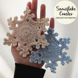 Könnte beinhalten: Zwei handgefertigte gehäkelte Schneeflocken-Untersetzer, einer hellrosa und einer hellblau, werden in einer Hand gehalten. Die Untersetzer sind kreisförmig mit aufwendigen, durchbrochenen Designs. Der Text auf dem Bild lautet "Snowflake Coaster CROCHET PATTERN PDF."
