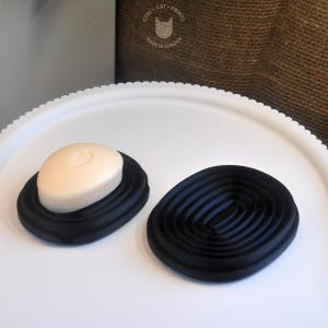 May include: Two black soap dishes, one holding a light beige oval soap. The soap dish has a spiral design. The soap has a small design on it. The text "CTRL CAT PRINTS MADE IN LONDON" is visible in the background.