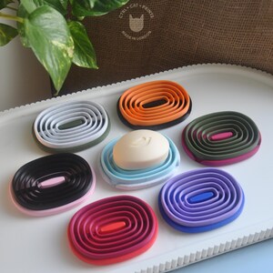 May include: A selection of colourful, oval-shaped soap dishes in shades like orange, black, pink, purple, and green. One dish holds a bar of white soap. The dishes are arranged on a white tray. The text "CTRL + CAT + PRINTS" and "MADE IN LONDON" is visible.