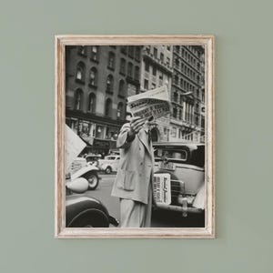 May include: A black and white framed photograph depicting a person in a double-breasted suit holding a newspaper titled Social Justice in front of their face. The scene features vintage cars and city buildings in the background of a street.
