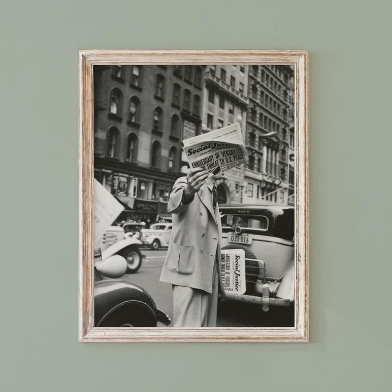 May include: A black and white framed photograph depicting a person in a double-breasted suit holding a newspaper titled Social Justice in front of their face. The scene features vintage cars and city buildings in the background of a street.