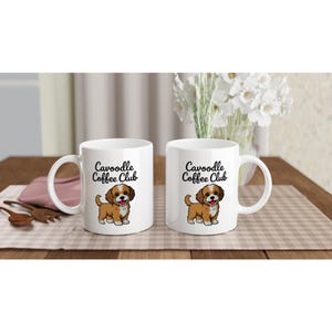 May include: Two white ceramic coffee mugs. Each mug features the text "Cavoodle Coffee Club" and a cartoon Cavapoo dog illustration. The mugs have a curved handle and are on a checkered surface.