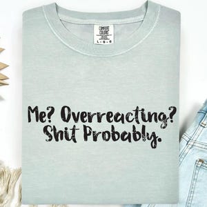 May include: A light sage green crew neck t-shirt featuring the black distressed text "Me? Overreacting? Shit Probably." printed on the front. The shirt has a Comfort Colors brand label at the inner neckline indicating a size large.