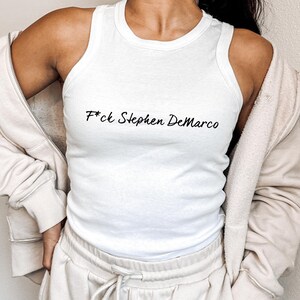 May include: White ribbed vest top featuring the text "F*ck Stephen DeMarco" in black script. The top is paired with a beige zip-up hoodie and matching joggers. The outfit has a relaxed, casual style.