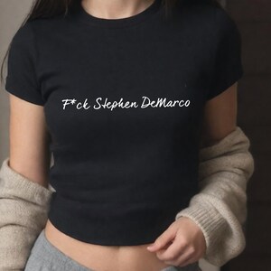 May include: Black crew neck t-shirt with the text "F*ck Stephen DeMarco" in white script. The shirt is short-sleeved and appears to be made of a soft material. A beige cardigan is draped over the shoulders.