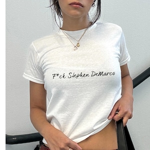 May include: White crew neck t-shirt featuring the black text "F*ck Stephen DeMarco". The short-sleeved top is a casual style.